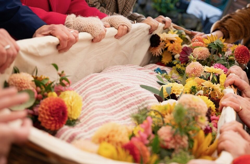 Many hands holding the edge of a woven coffin, filled with beautiful cloth and flowers